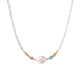 Delicate white beaded necklace with baroque pearl center, gold accents, and coral and aqua gemstones on cream background.